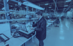 Technician working on electronics assembly in a modern manufacturing facility with production equipment and workstations in the background.