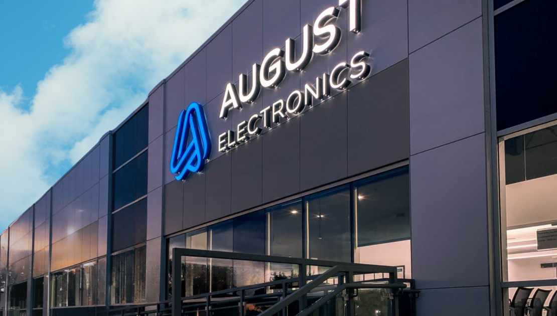 Exterior of August Electronics building with illuminated logo, showcasing modern facility for Electronics Manufacturing Services (EMS) site visits.