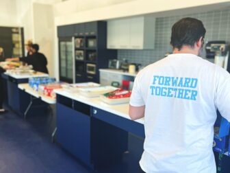 A person wearing a white t-shirt with the text 'FORWARD TOGETHER' printed on the back, standing in a kitchen area with various food items and drinks laid out on counters. Other people are seen in the background preparing or serving food.