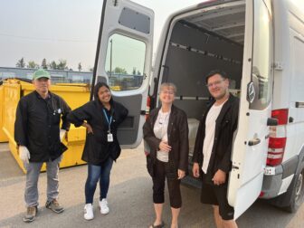 Four people standing next to an open white van, with two large yellow bins in the background. They are all wearing black jackets. The person on the far left is wearing a green cap and gloves, the second person is smiling with hands on hips and wearing an ID badge, the third person is smiling with hands folded, and the fourth person is standing inside the van, smiling and wearing glasses.