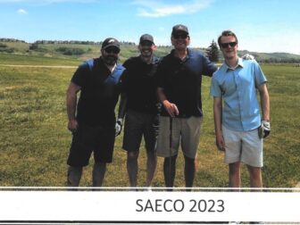 Four men standing together on a golf course, smiling at the camera, with the text 'SAECO 2023' below the photo. They are dressed in casual golf attire, and the background features a grassy field and a clear blue sky.